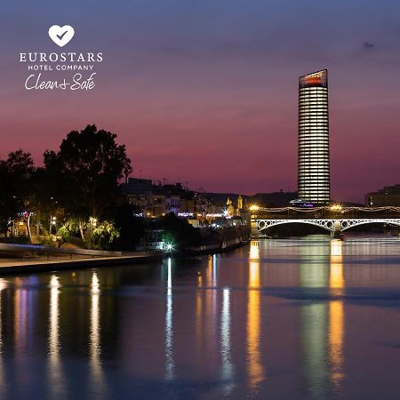 Eurostars Torre Sevilla Esterno foto The photo depicts a serene riverside view at dusk. A tall, modern skyscraper with illuminated windows stands in the background against a twilight sky transitioning from orange to deep blue. On the left, there are trees and buildings that add to the u