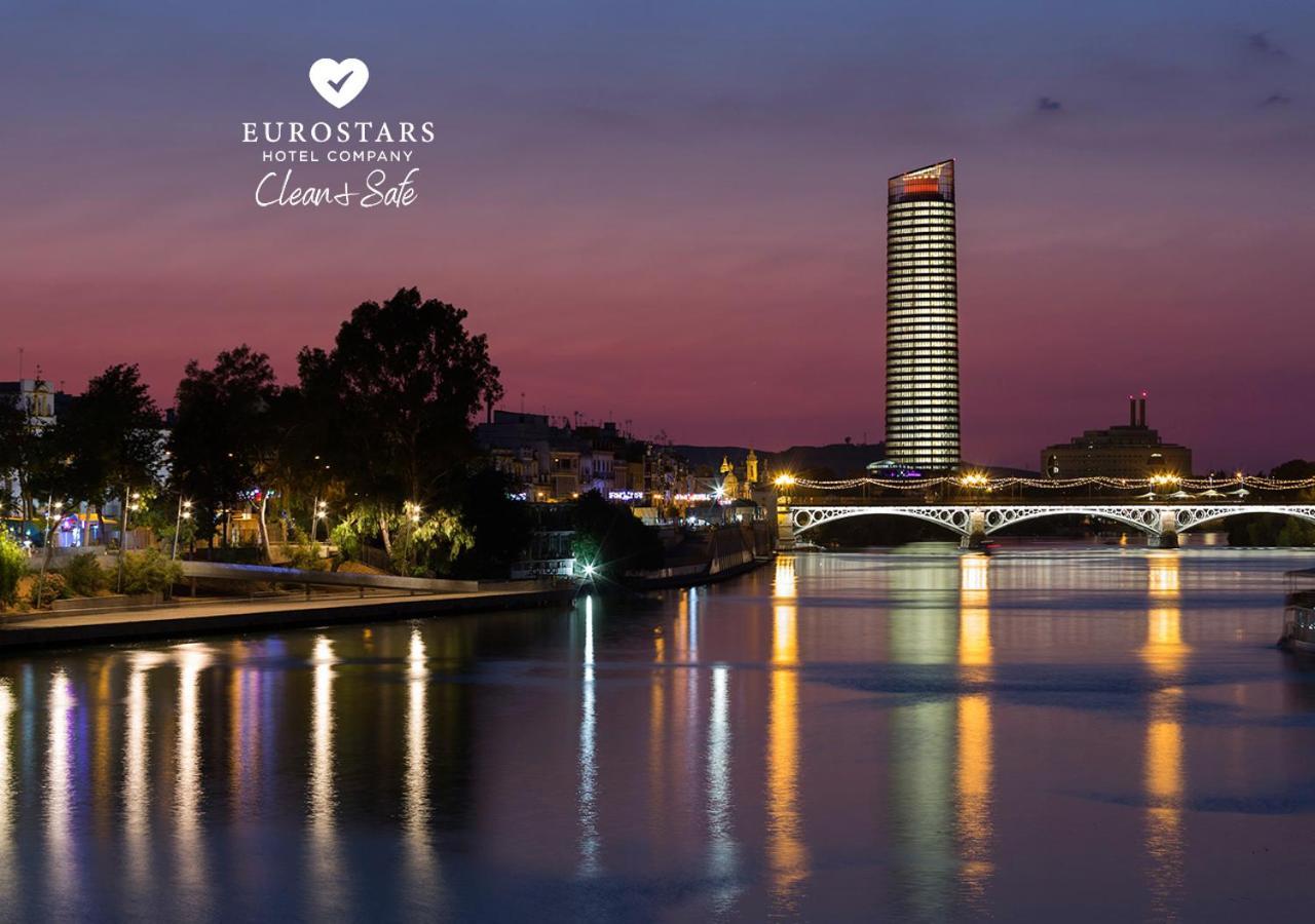 Eurostars Torre Sevilla Esterno foto The photo depicts a serene riverside view at dusk. A tall, modern skyscraper with illuminated windows stands in the background against a twilight sky transitioning from orange to deep blue. On the left, there are trees and buildings that add to the u