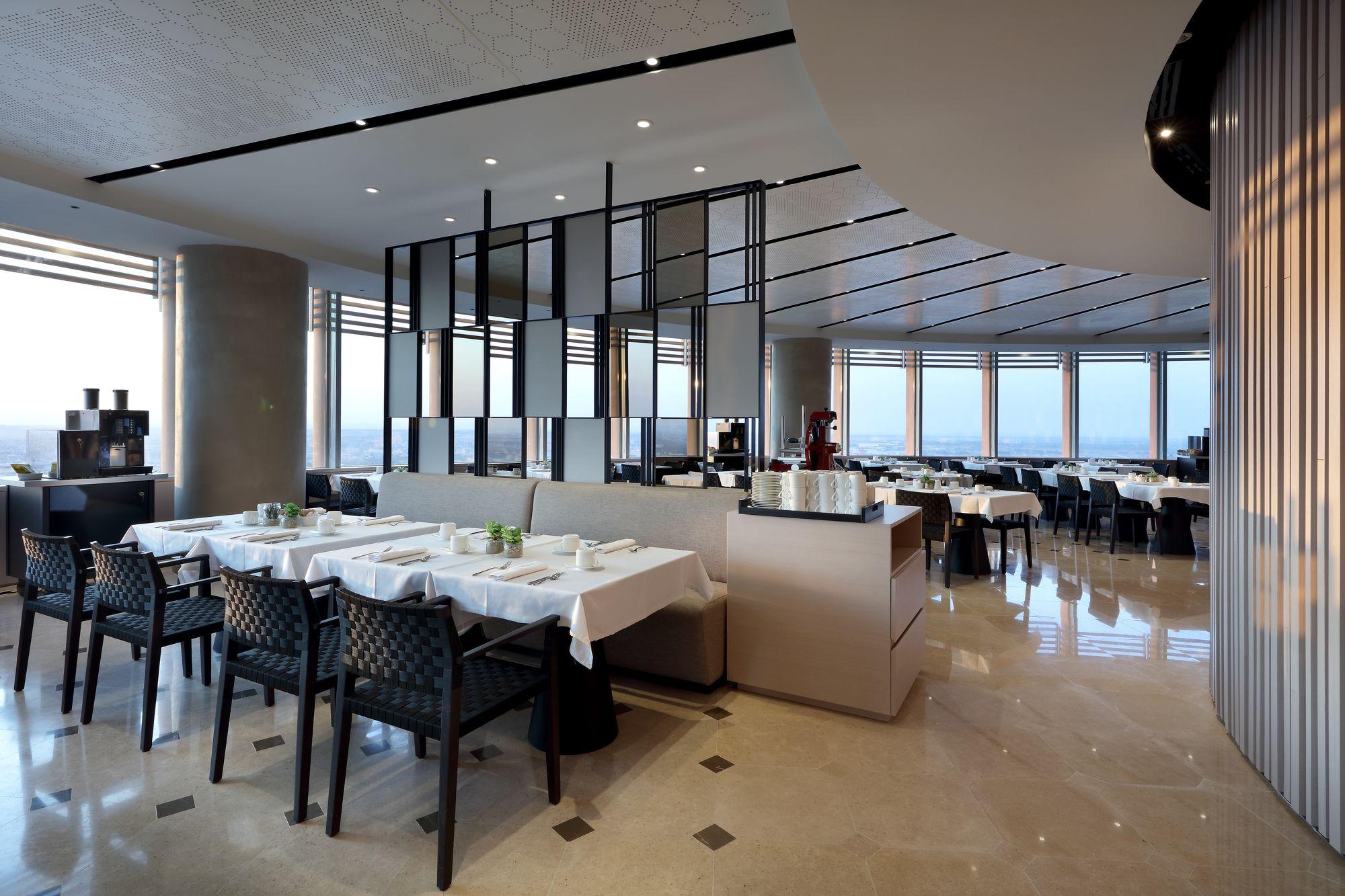 Eurostars Torre Sevilla Esterno foto The photo shows the interior of a modern restaurant. The design features a curved layout with elegant decor. There are several tables set with white tablecloths and black chairs. Large windows provide a view of the outside, allowing natural light to 
