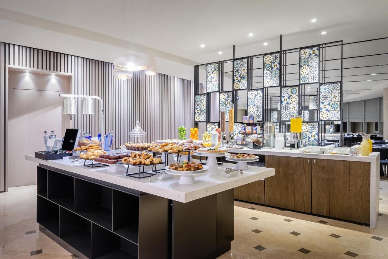 Eurostars Torre Sevilla Esterno foto The photo shows a modern buffet setup in a dining area. There is a stylish display featuring various food items, including pastries and desserts, arranged on a central island with multiple tiers. Behind the food spread, there is a decorative partitio