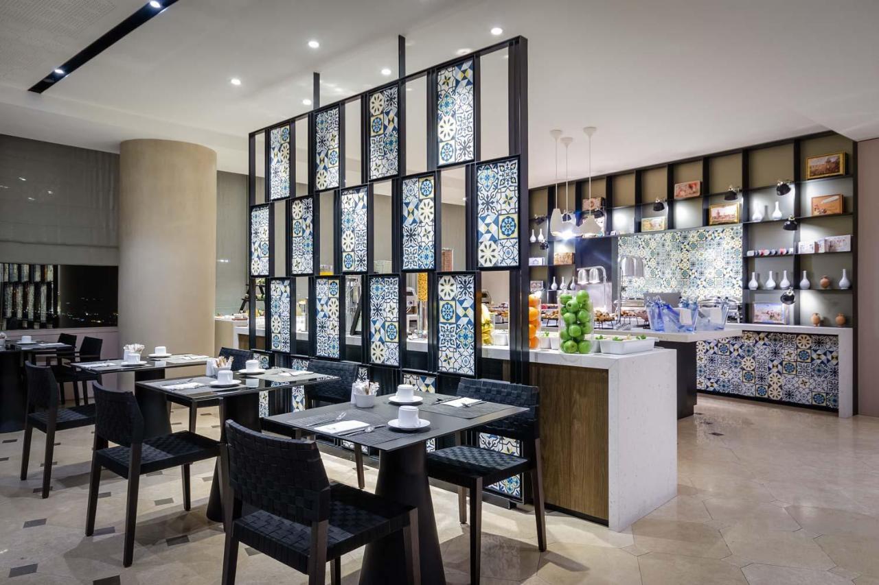 Eurostars Torre Sevilla Esterno foto The photo shows a restaurant interior featuring a modern design. There are several dining tables set for meal service, each with black chairs and neatly arranged tableware, including cups and plates. A decorative partition with colorful tile work sep