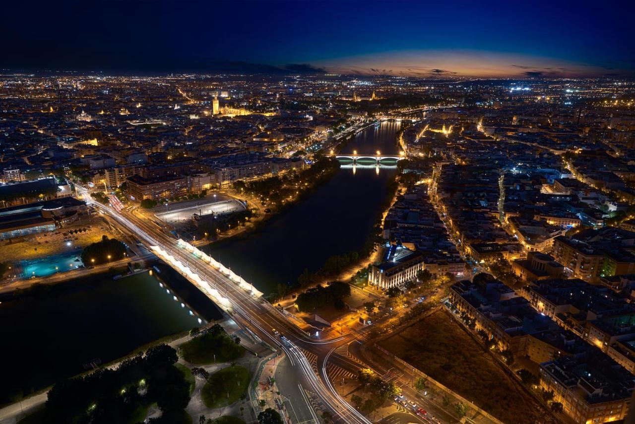 Eurostars Torre Sevilla Esterno foto The photo shows a panoramic view of a city at dusk, with a blend of natural and artificial light illuminating the landscape. A river runs through the scene, reflecting the city lights. There are several bridges crossing the river, and the buildings a