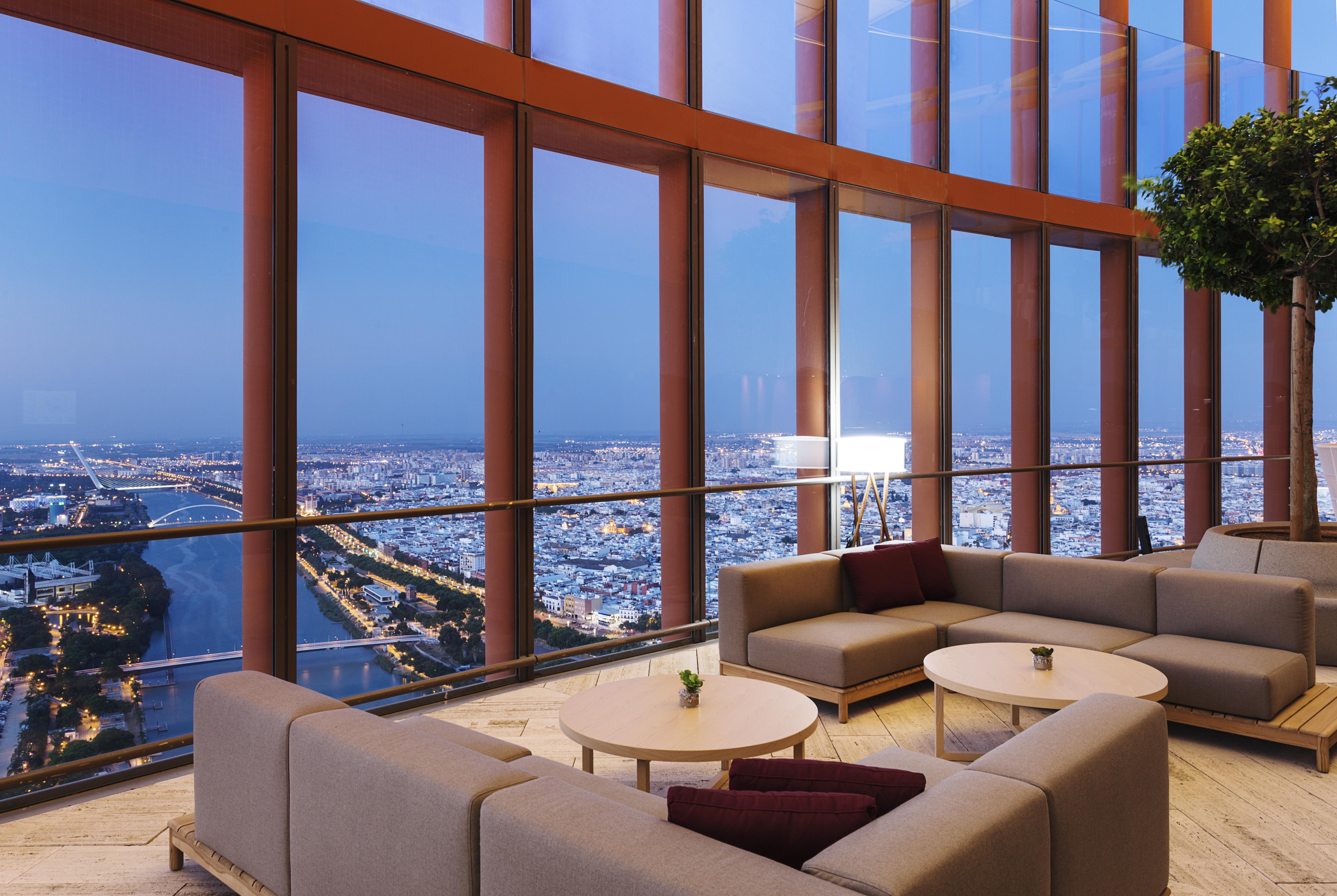 Eurostars Torre Sevilla Esterno foto The photo shows a modern, stylish interior space with large windows providing a panoramic view of a cityscape at dusk. The room features a comfortable seating arrangement with light-colored sofas and a couple of small tables. There are some decorativ