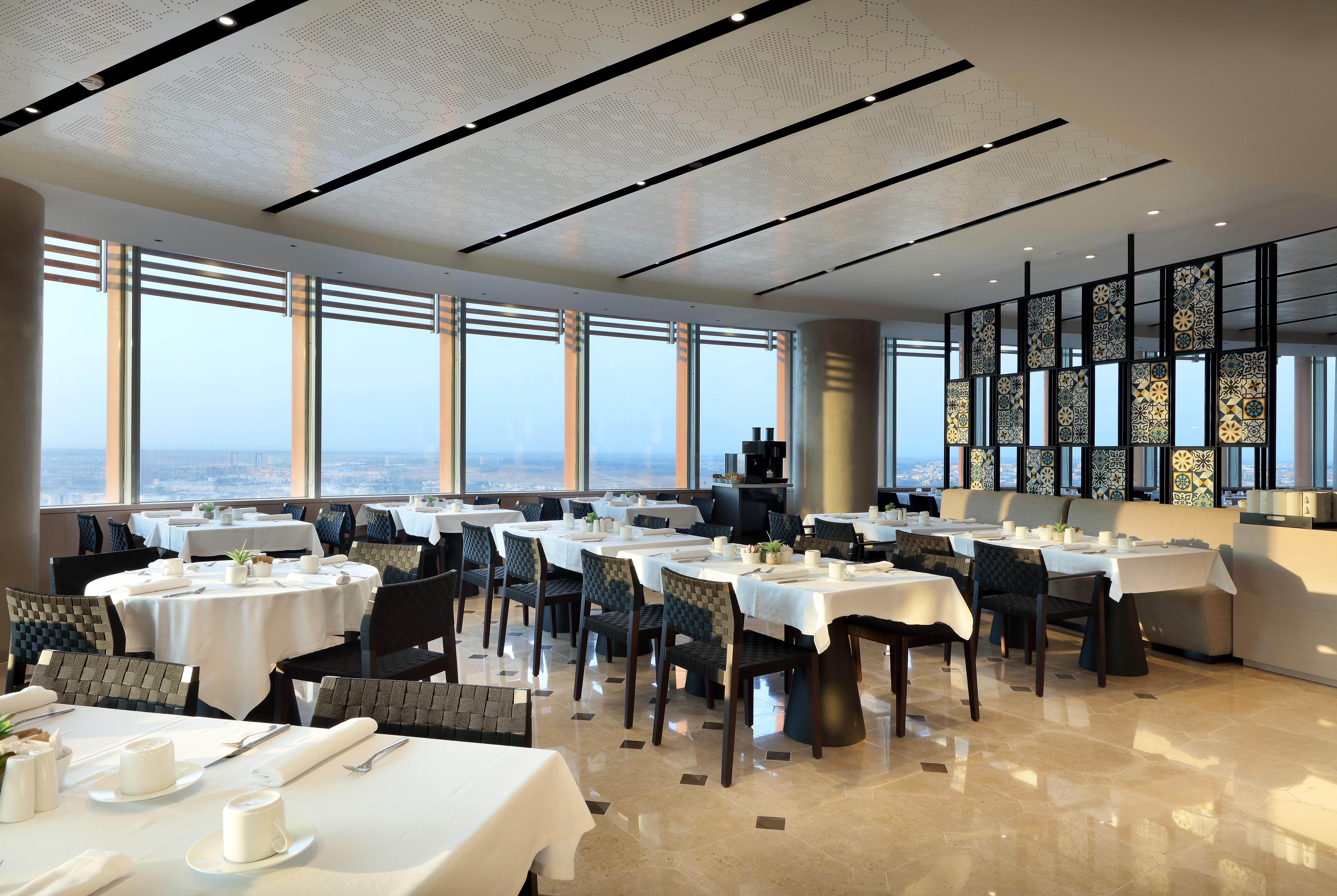 Eurostars Torre Sevilla Esterno foto The photo shows the interior of a modern restaurant. The dining area features several tables covered with white tablecloths, each set with utensils and cups. There are black chairs around the tables. The space is well-lit by large windows that likely