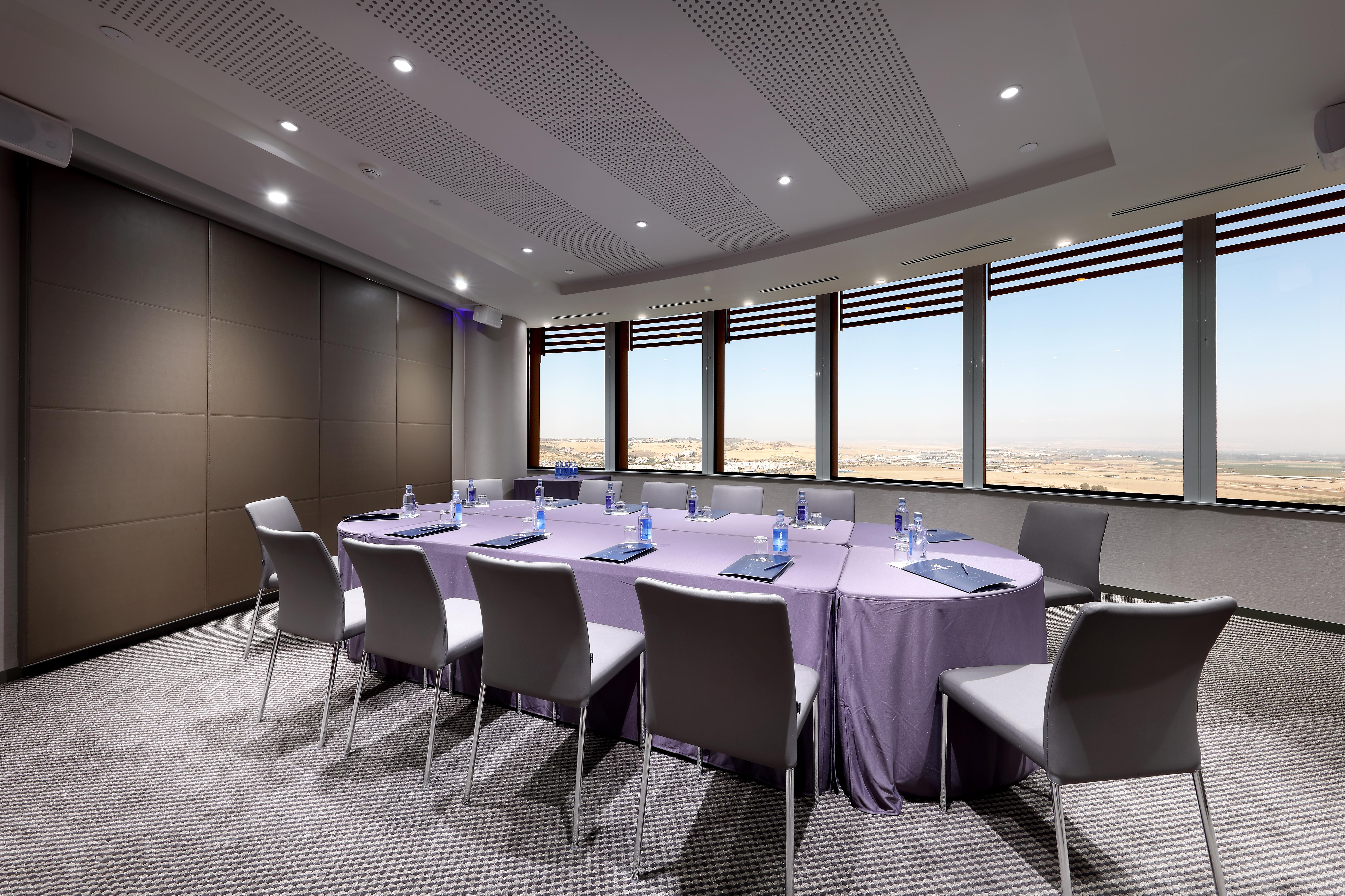 Eurostars Torre Sevilla Esterno foto The photo shows a modern conference room designed for meetings. In the center, there is a round table covered with a purple tablecloth, surrounded by several chairs. Each chair has a light gray design. On the table, there are bottles of water and not