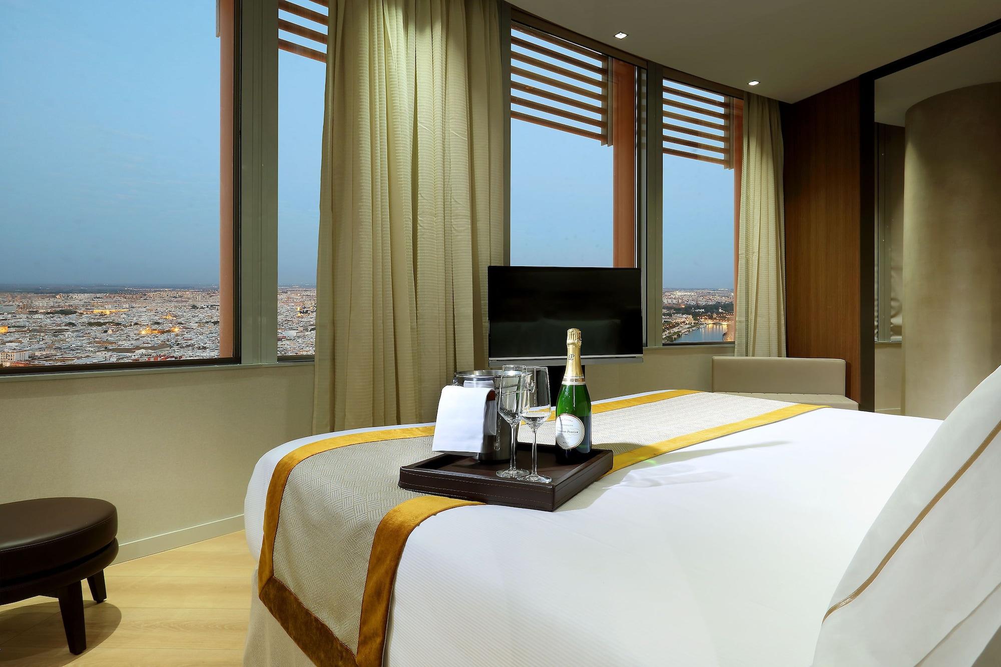 Eurostars Torre Sevilla Esterno foto The photo shows a modern hotel room featuring a large bed covered with a light-colored bedspread that has a gold decorative stripe. On a tray in the center of the bed, there are two champagne flutes, a bottle of champagne, and a small bowl of snacks.
