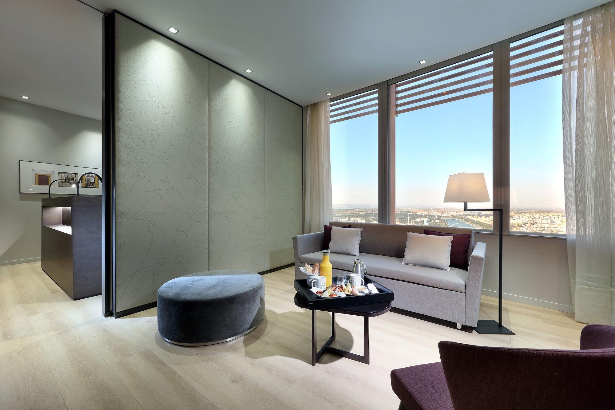 Eurostars Torre Sevilla Esterno foto The photo depicts a modern, stylish interior of a hotel room or suite. It features a comfortable seating area with a grey sofa, a round ottoman, and a small table that appears to hold refreshments. Large windows allow natural light to fill the space,