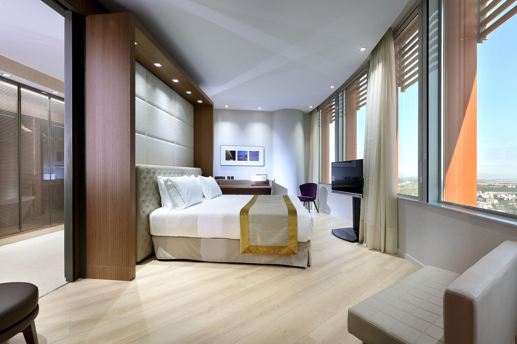 Eurostars Torre Sevilla Esterno foto The photo shows a modern hotel room featuring a large bed with a light-colored headboard. The decor is minimalist, with a neutral color palette and wooden accents. A small seating area is located next to the bed, and there is a desk with a chair. A t