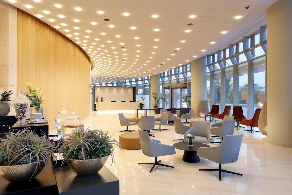 Eurostars Torre Sevilla Esterno foto The photo shows a modern, well-lit lobby or lounge area. The design features smooth lines and a contemporary aesthetic, with a curved wooden wall and large windows allowing natural light to fill the space. There are stylish seating arrangements, incl