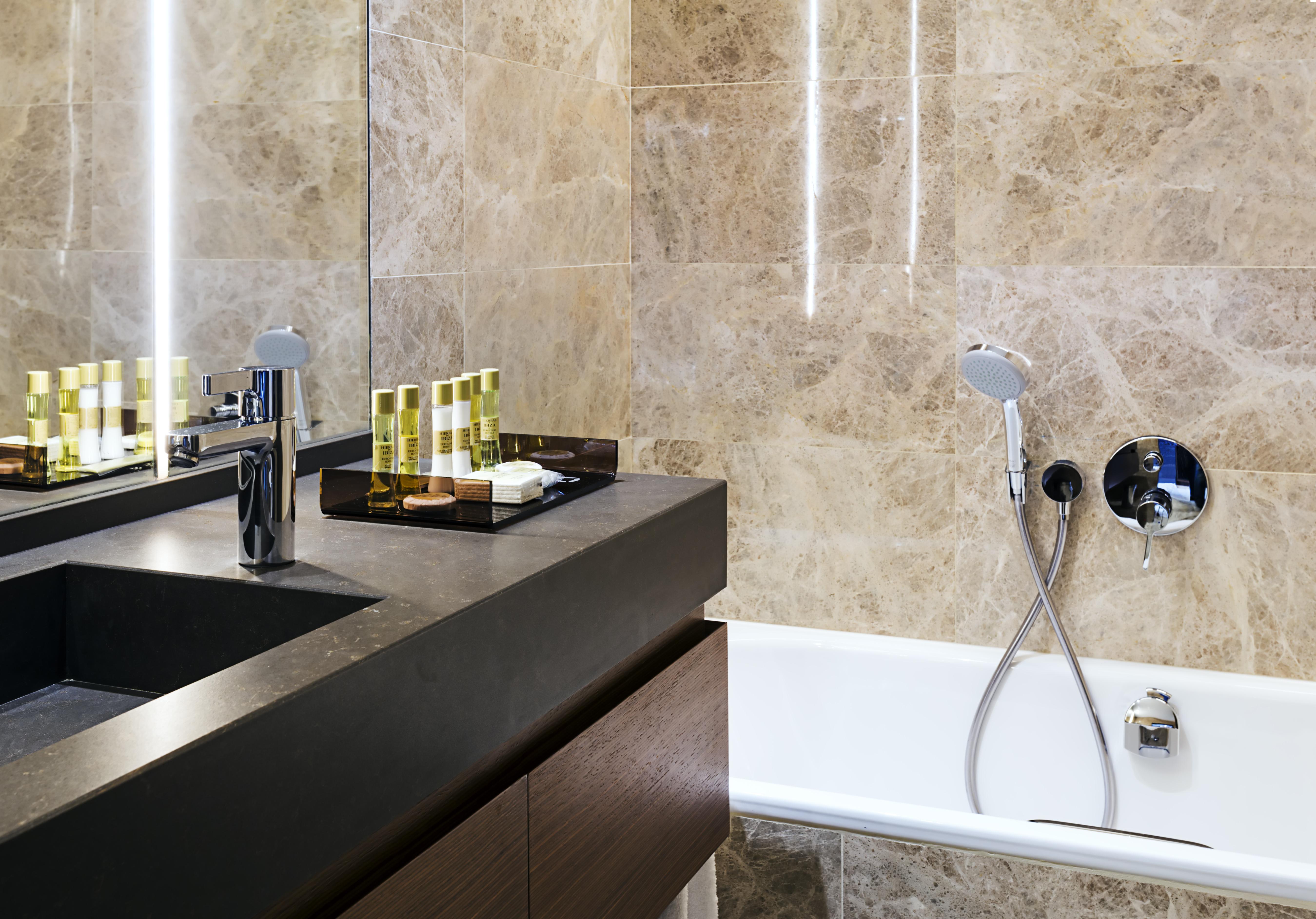 Eurostars Torre Sevilla Esterno foto The photo shows a modern bathroom featuring elegant design elements. There is a sleek, dark countertop with a rectangular sink and a modern faucet. On the countertop, there are several small bottles, likely containing toiletries, arranged neatly on a
