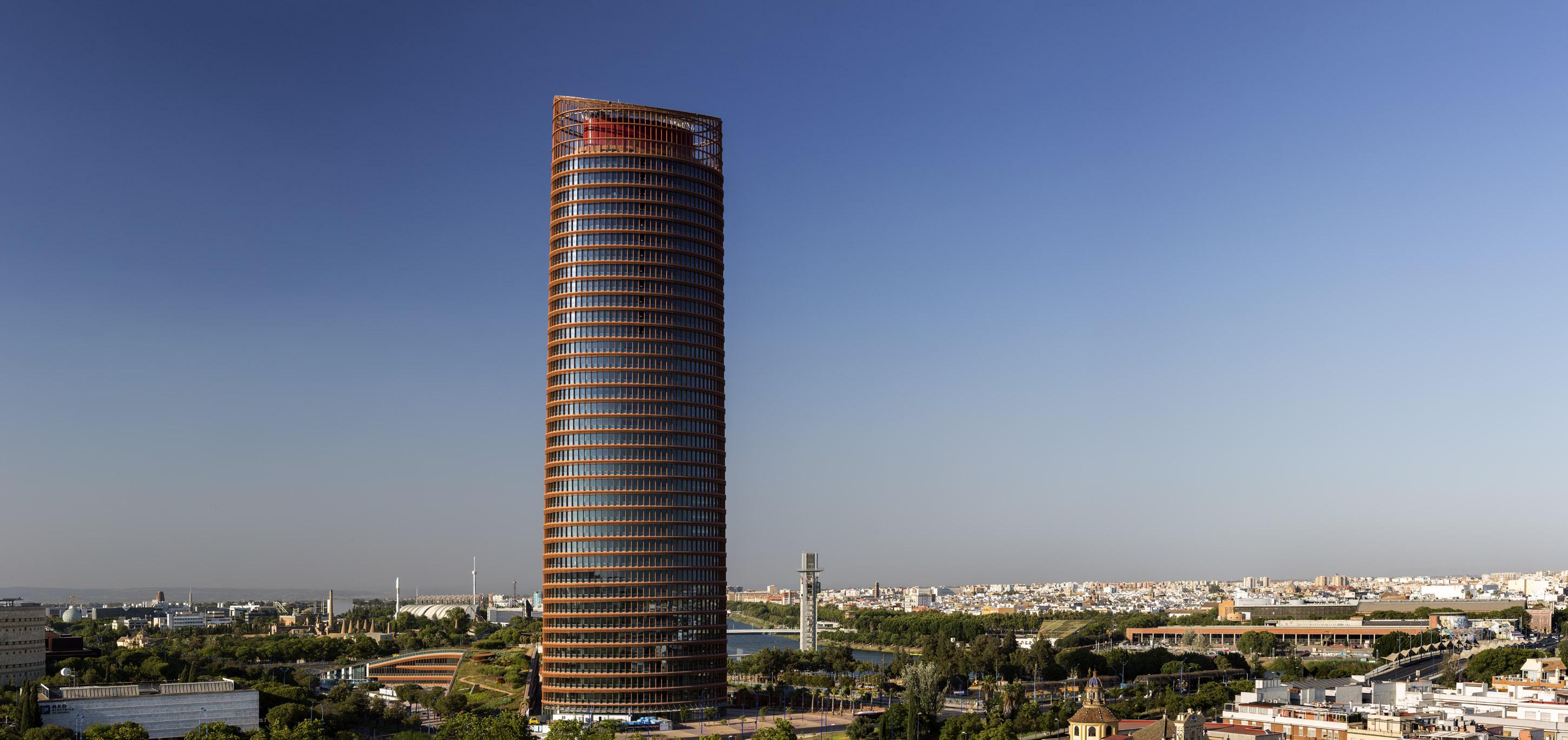 Eurostars Torre Sevilla Esterno foto The photo showcases a tall, modern skyscraper with a cylindrical shape and a distinctive facade featuring a combination of glass and metal elements. The building is surrounded by greenery and urban landscapes, with other structures visible in the bac