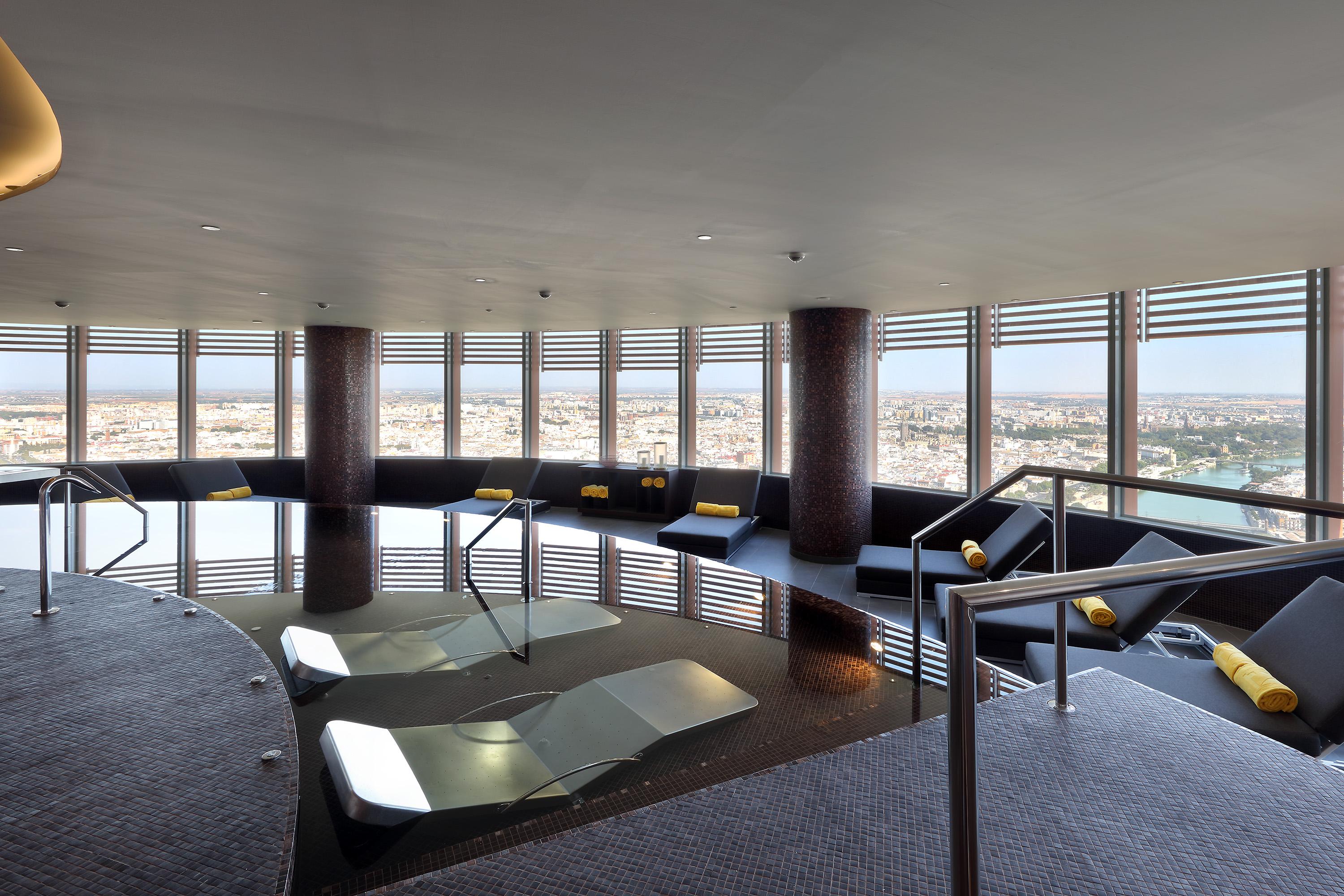 Eurostars Torre Sevilla Esterno foto The photo depicts an elegant spa or wellness area with a large, circular pool surrounded by comfortable seating. The space features large windows that offer a panoramic view of the city outside, allowing natural light to illuminate the room. The inte