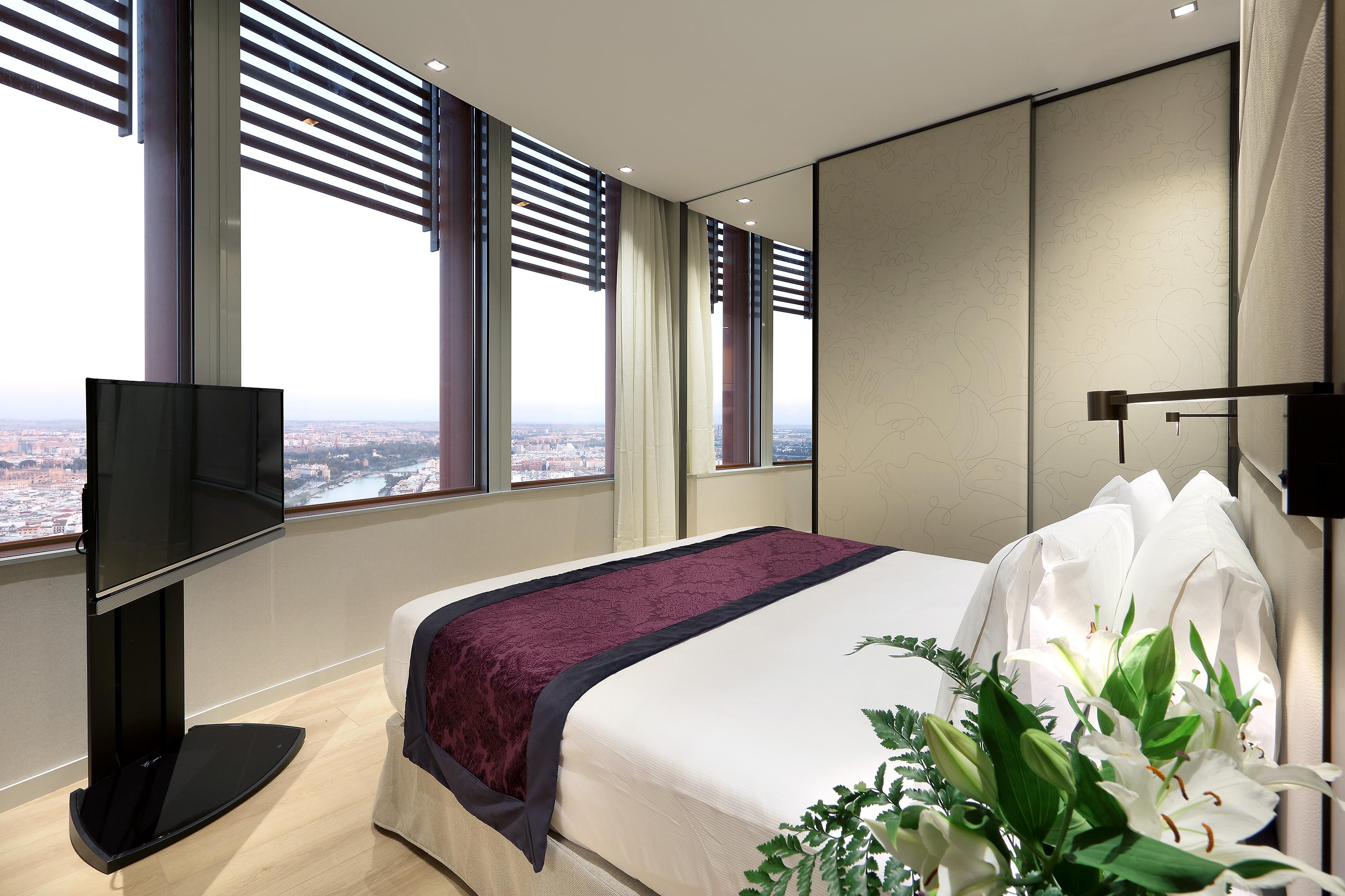 Eurostars Torre Sevilla Esterno foto The image shows a modern hotel room featuring a large bed with a dark purple bedspread and white pillows. The room has floor-to-ceiling windows with horizontal blinds, providing a panoramic view of the city outside. A flat-screen television is mounte
