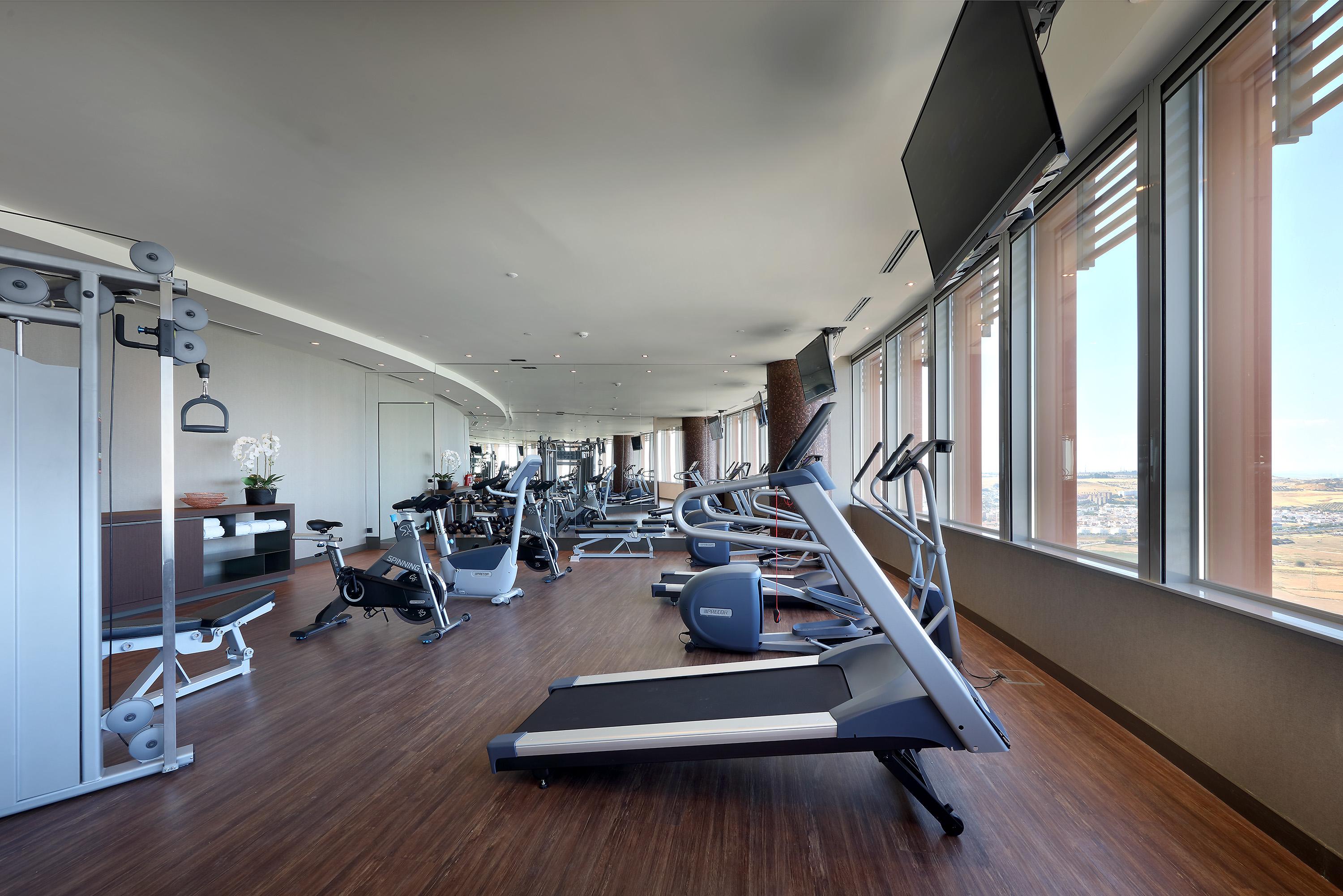Eurostars Torre Sevilla Esterno foto The photo shows a modern gym facility with large windows allowing natural light to enter. There are various exercise machines, including treadmills, stationary bikes, and strength training equipment. The flooring appears to be wood, contributing to a
