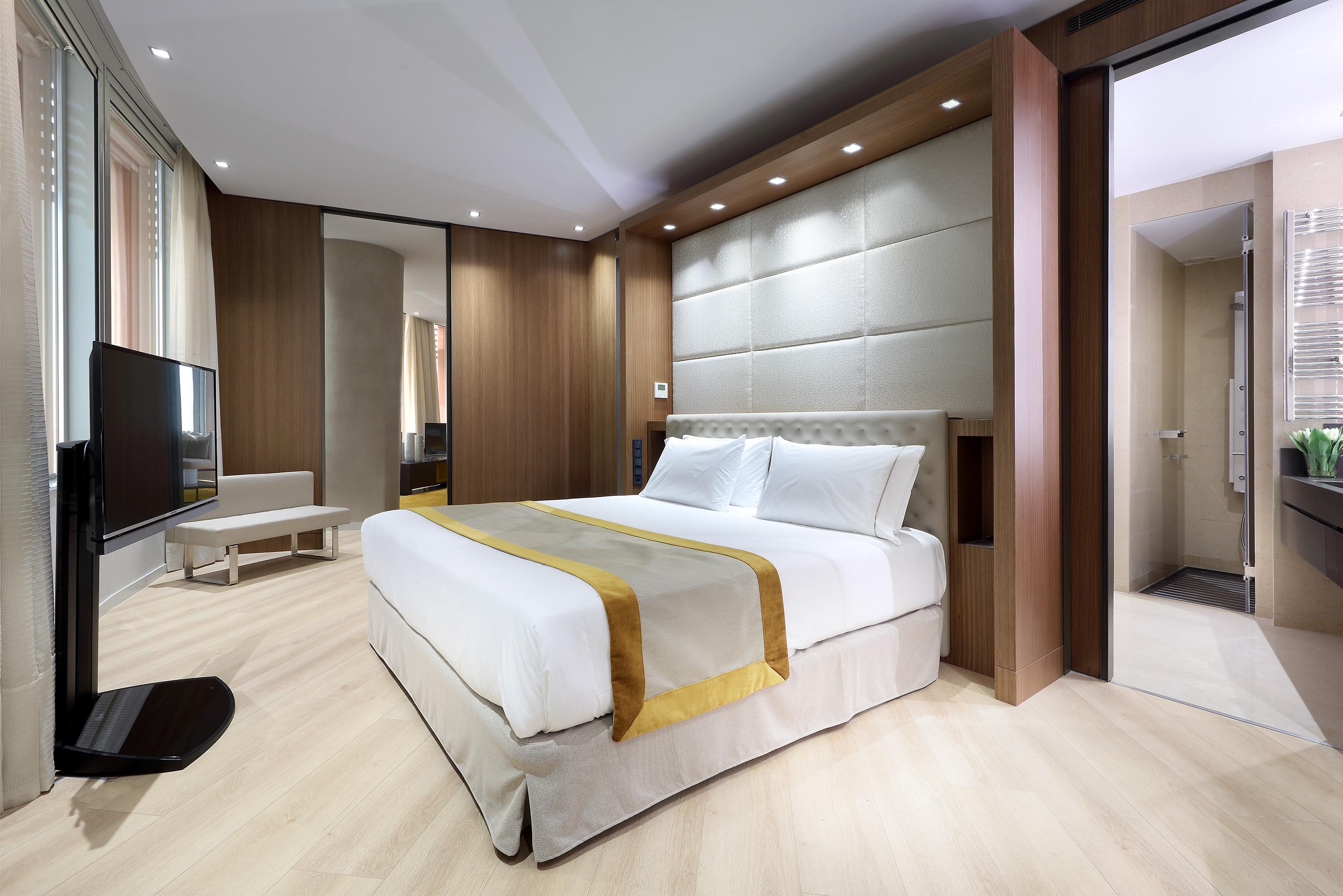 Eurostars Torre Sevilla Esterno foto The photo depicts a modern hotel room featuring a neatly made bed with a light-colored coverlet and a decorative gold stripe. The room has a contemporary aesthetic, characterized by wood paneling and soft lighting. A flat-screen TV is mounted on the 