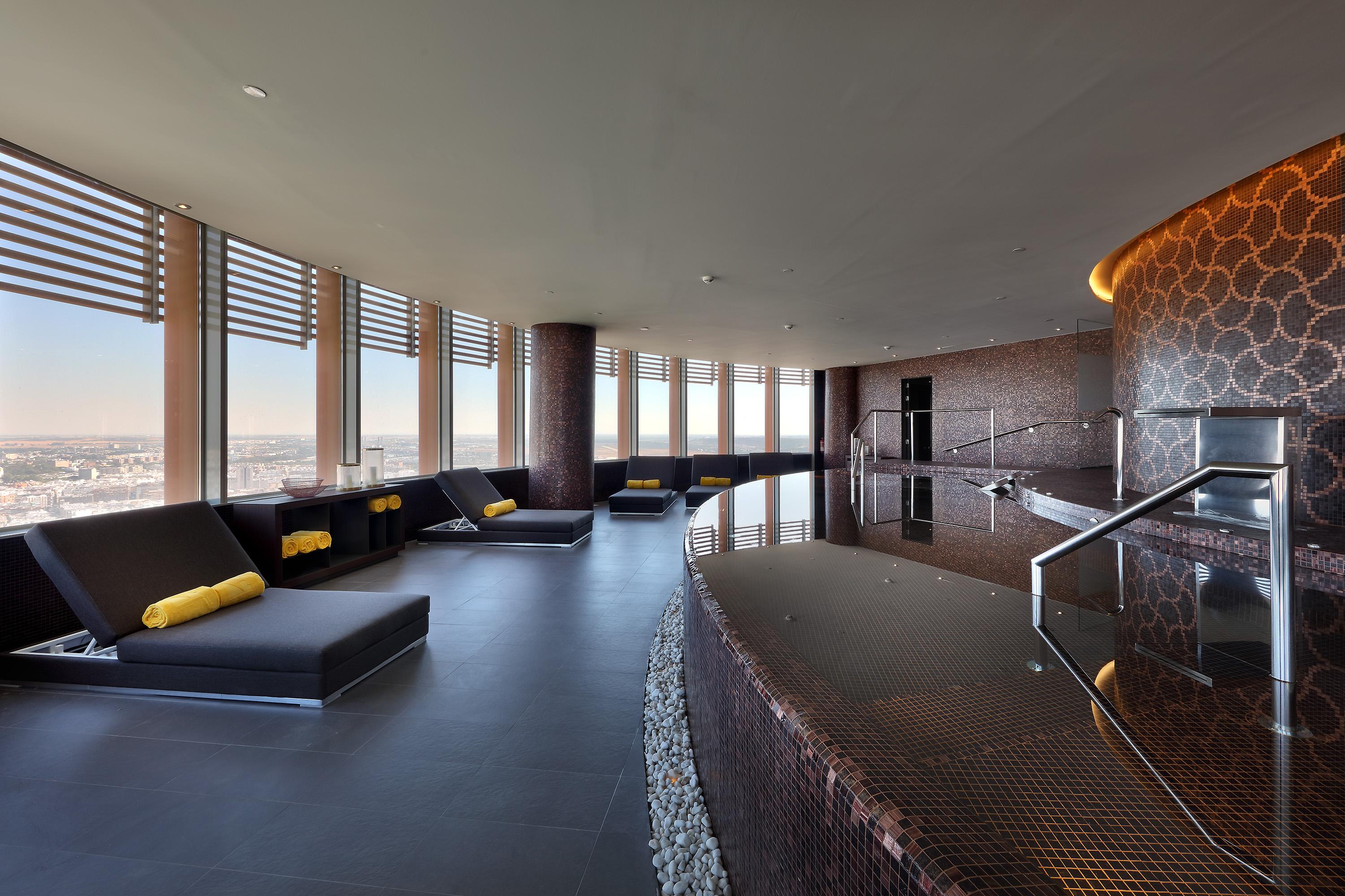 Eurostars Torre Sevilla Esterno foto The photo depicts a modern and elegant spa interior. It features a curved layout with large windows providing a panoramic view. The space includes several lounge chairs with yellow cushions, arranged in a relaxed manner. A sleek, dark-finished counte