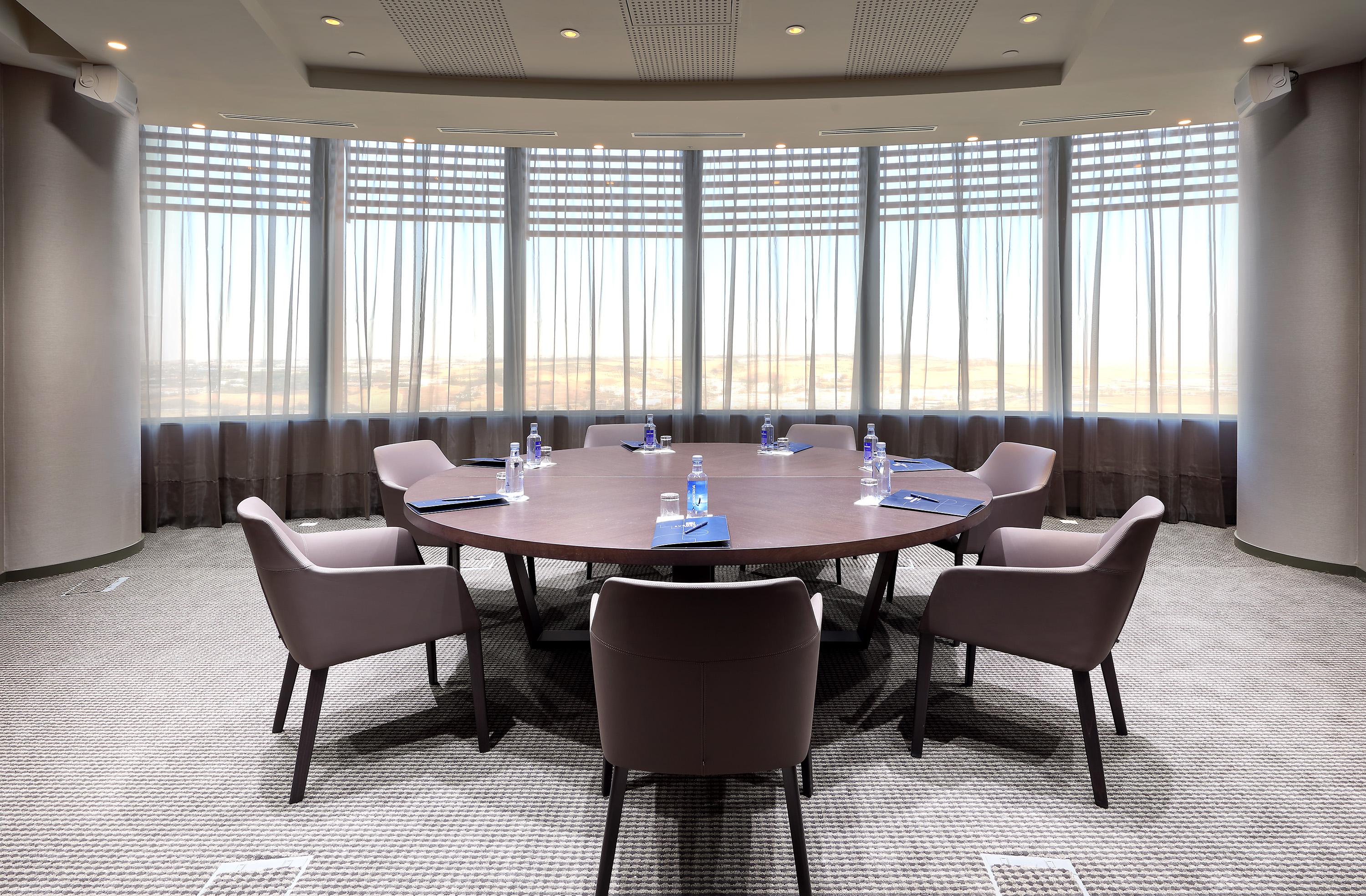 Eurostars Torre Sevilla Esterno foto The photo shows a conference room with a large round table at its center. The table is surrounded by several modern chairs. There are water bottles placed at each seat, and blue notepads or documents are also visible on the table. The room has large 