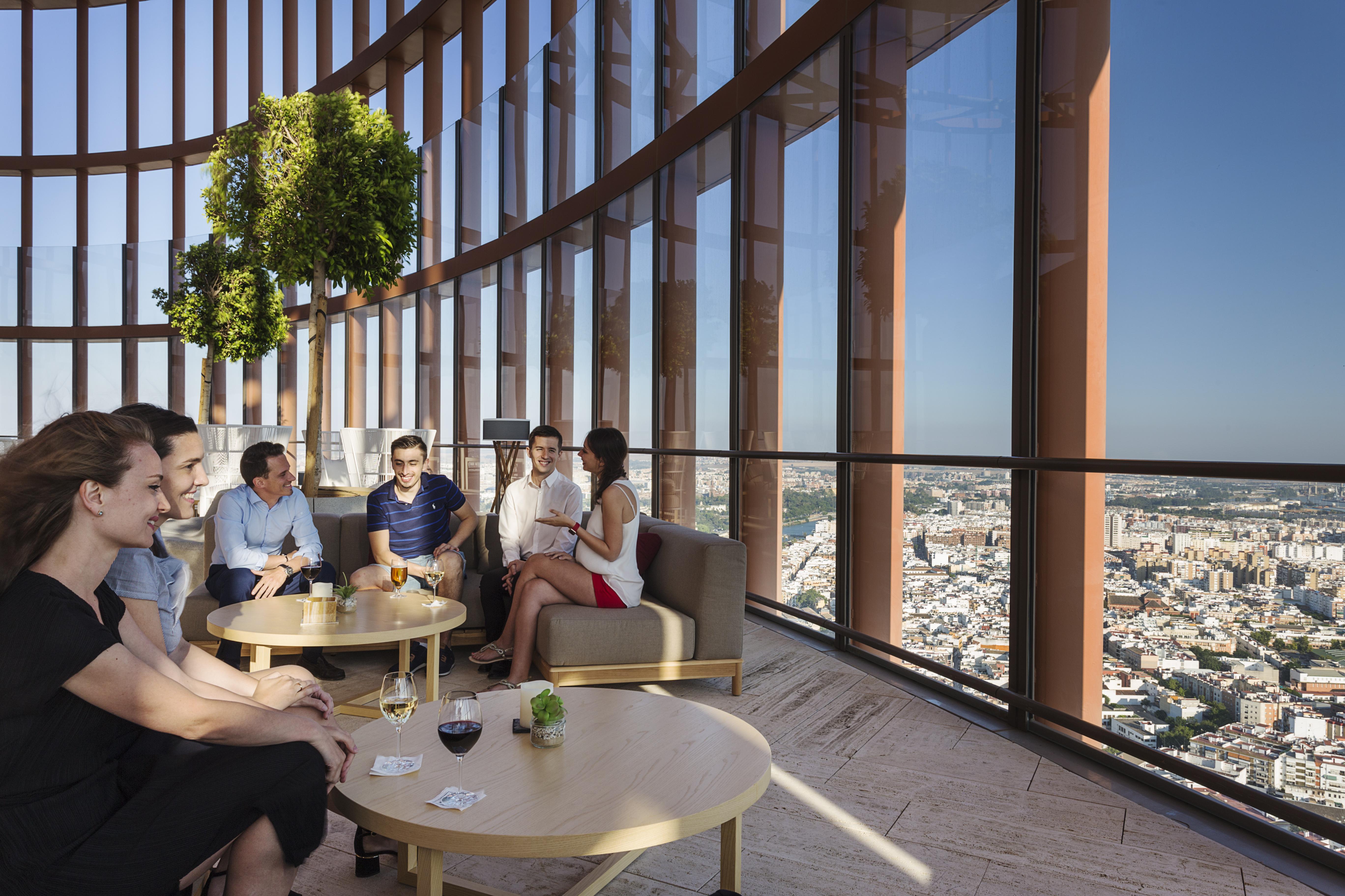 Eurostars Torre Sevilla Esterno foto The photo shows a group of people sitting in a modern, stylish lounge area with large windows that provide a panoramic view of a cityscape below. The setting appears to be high up in a building, possibly a skyscraper. There are several tables with dr