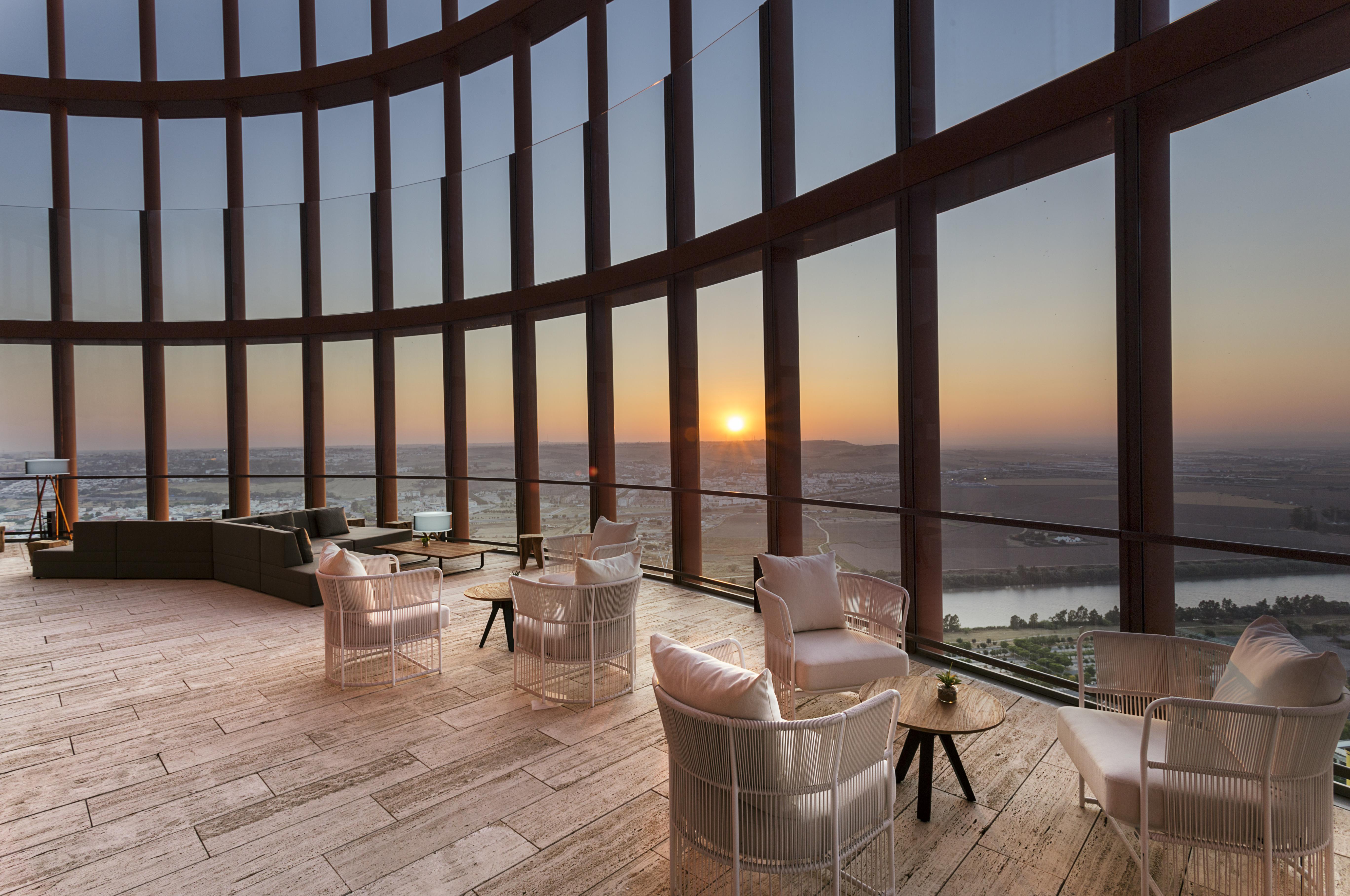 Eurostars Torre Sevilla Esterno foto The photo shows a spacious interior with large windows that provide a panoramic view of a sunset over a landscape. The room features modern furniture, including several white chairs and a small side table. The flooring appears to be made of light-col