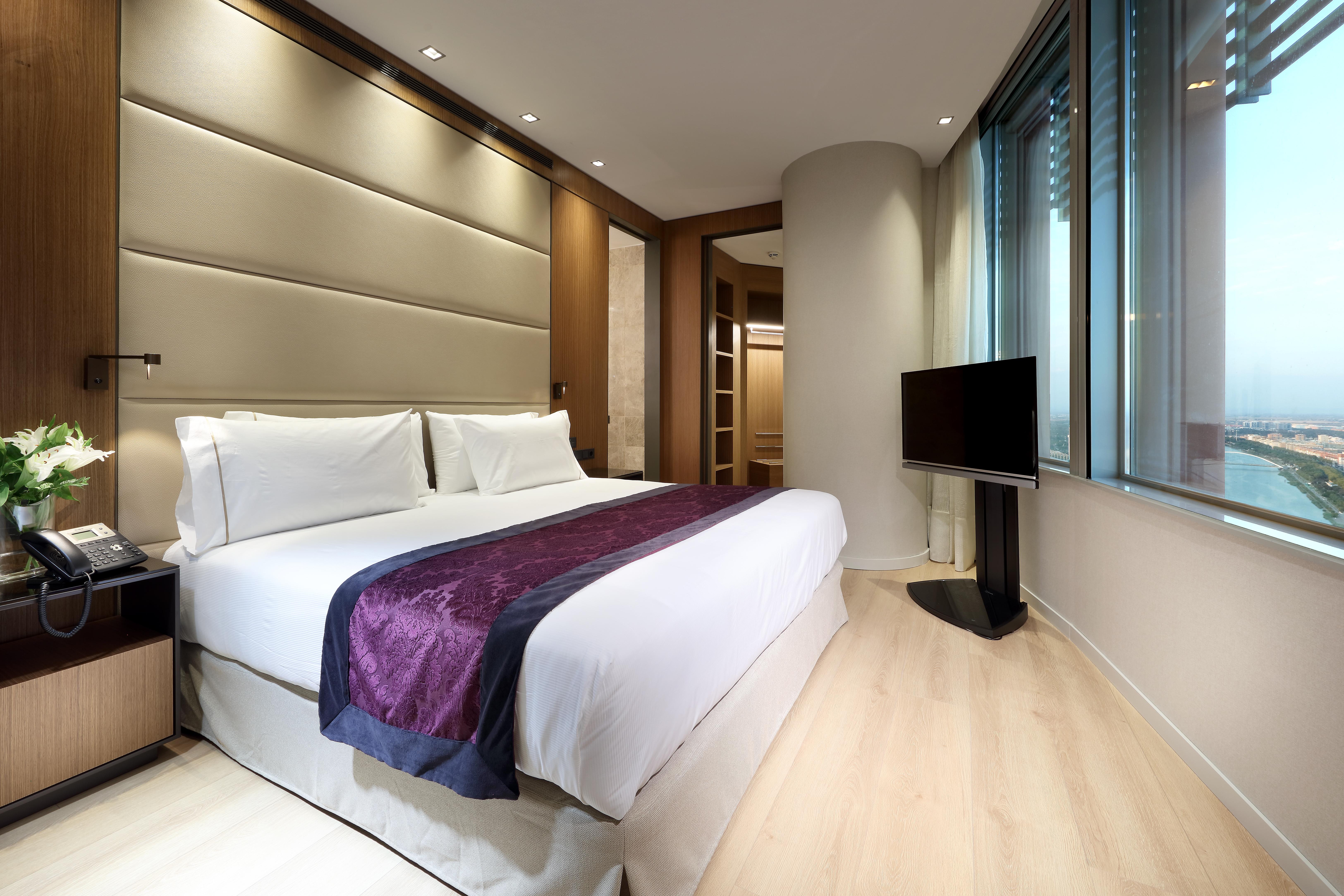 Eurostars Torre Sevilla Esterno foto The photo showcases a modern hotel room featuring a king-sized bed with white linens and a decorative purple accent. The room has wooden flooring and is well-lit with recessed lighting. Behind the bed, there is a plush headboard with a textured desig