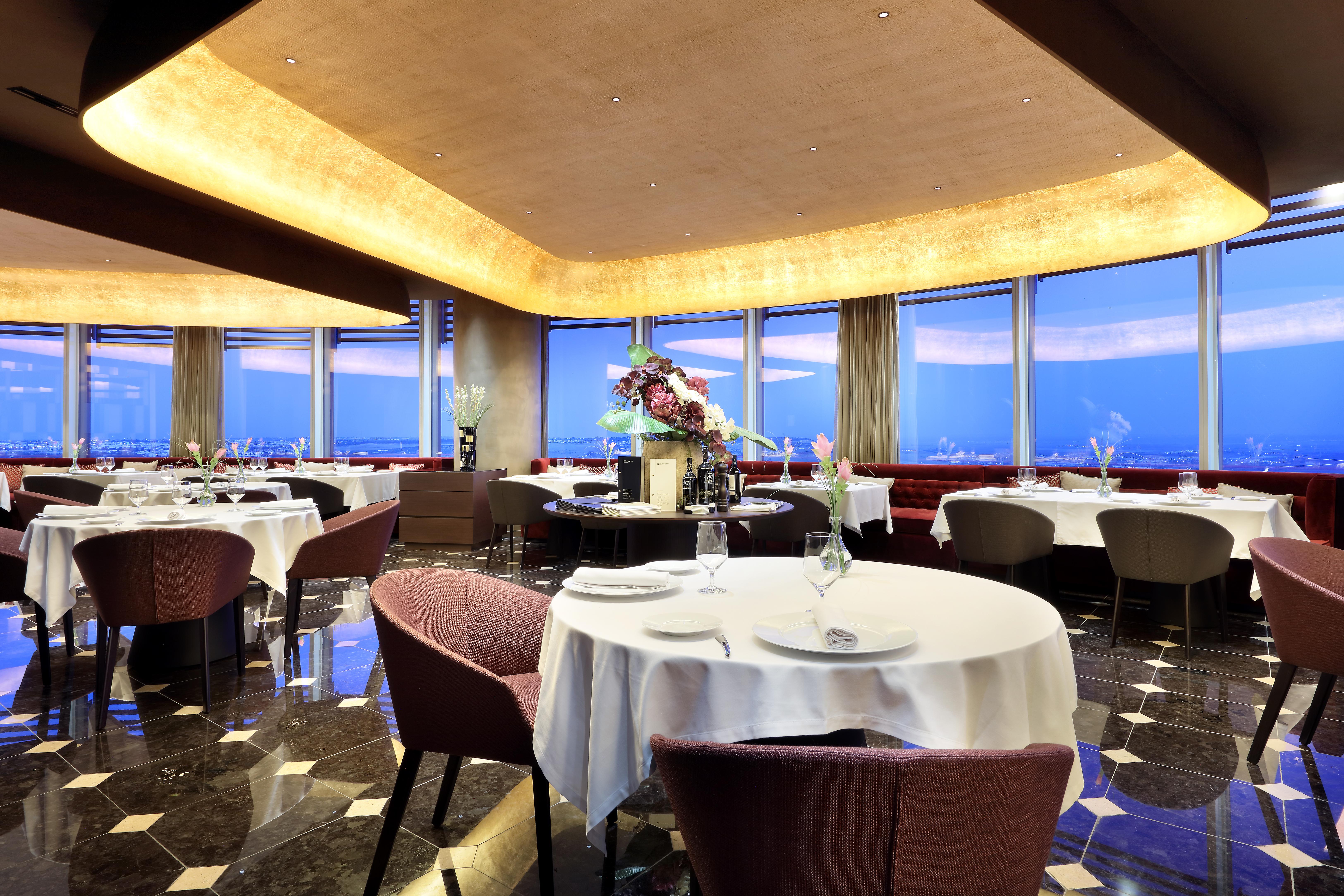 Eurostars Torre Sevilla Esterno foto The photo shows an elegant restaurant interior with a modern design. There are several round tables, each set with white tablecloths and place settings, including glasses and dishes. The seating consists of plush chairs that appear comfortable. The r