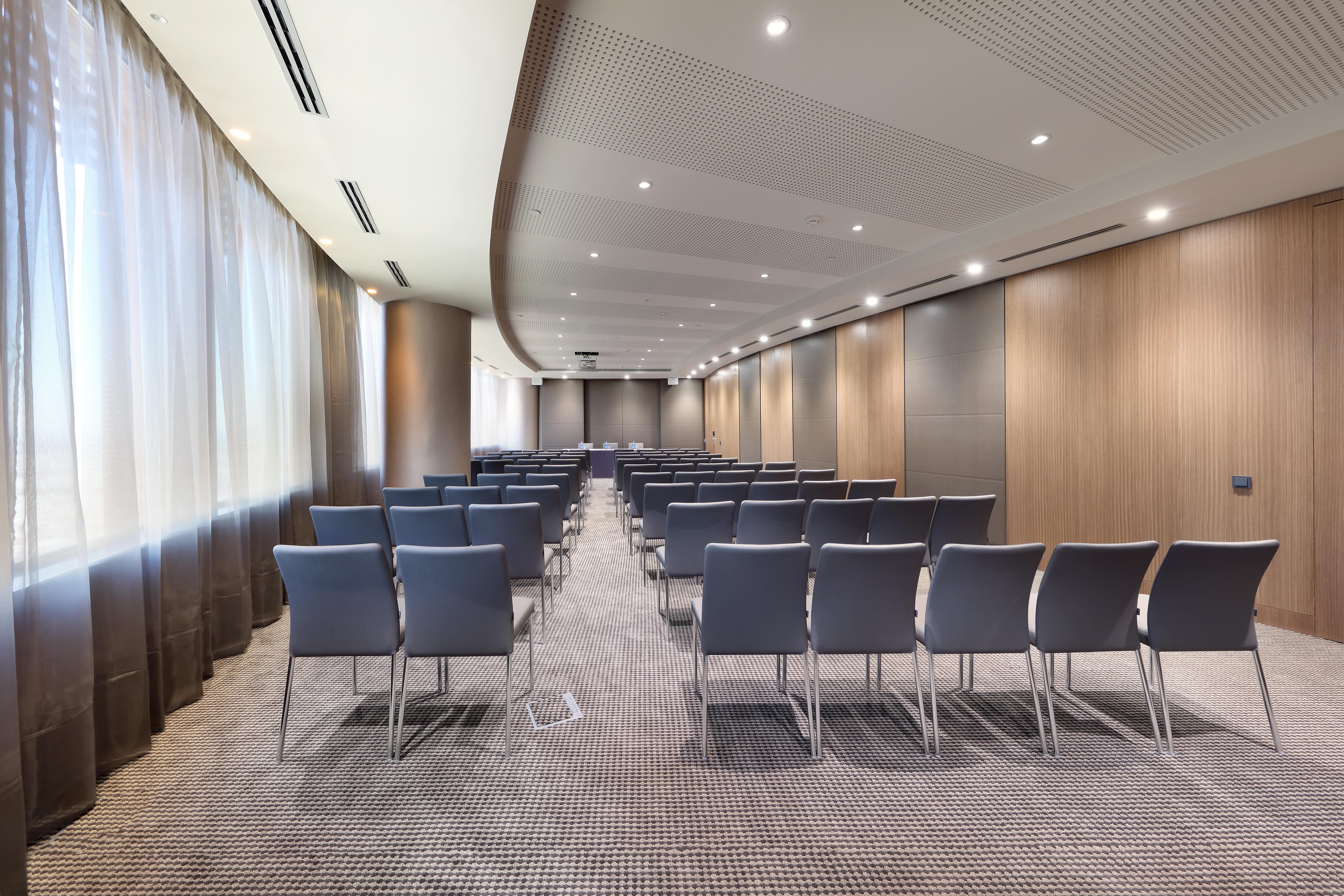Eurostars Torre Sevilla Esterno foto The photo shows a modern conference room with an elongated layout. There are rows of grey chairs arranged neatly facing a focal point at the front of the room, likely for a presentation or meeting. The walls are a combination of wooden paneling and n