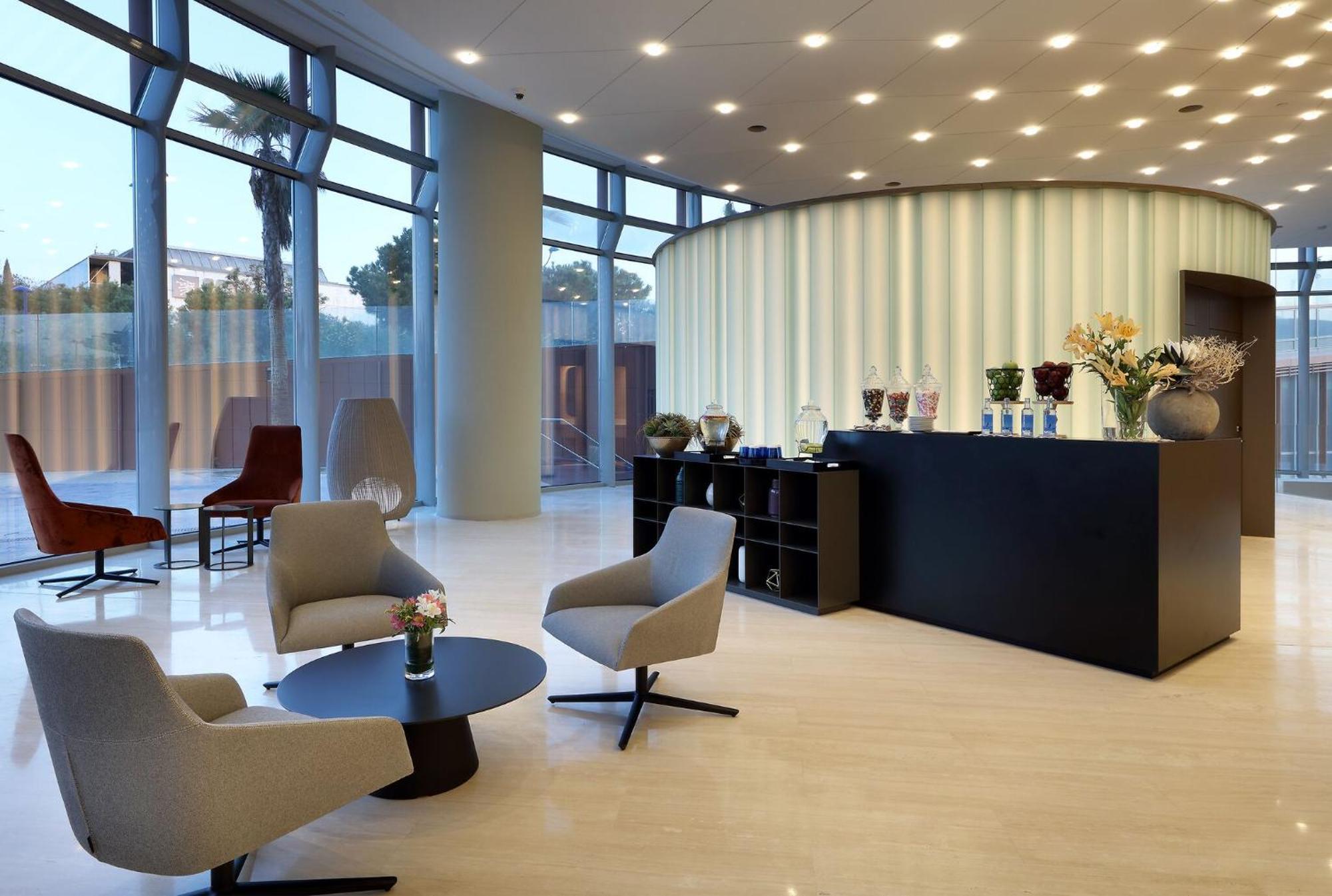 Eurostars Torre Sevilla Esterno foto The photo depicts a modern reception area. There is a sleek, rounded reception desk in the center against a light-colored wall with vertical stripes. Behind the desk, various decorative items and flowers are displayed. In front of the desk, there are