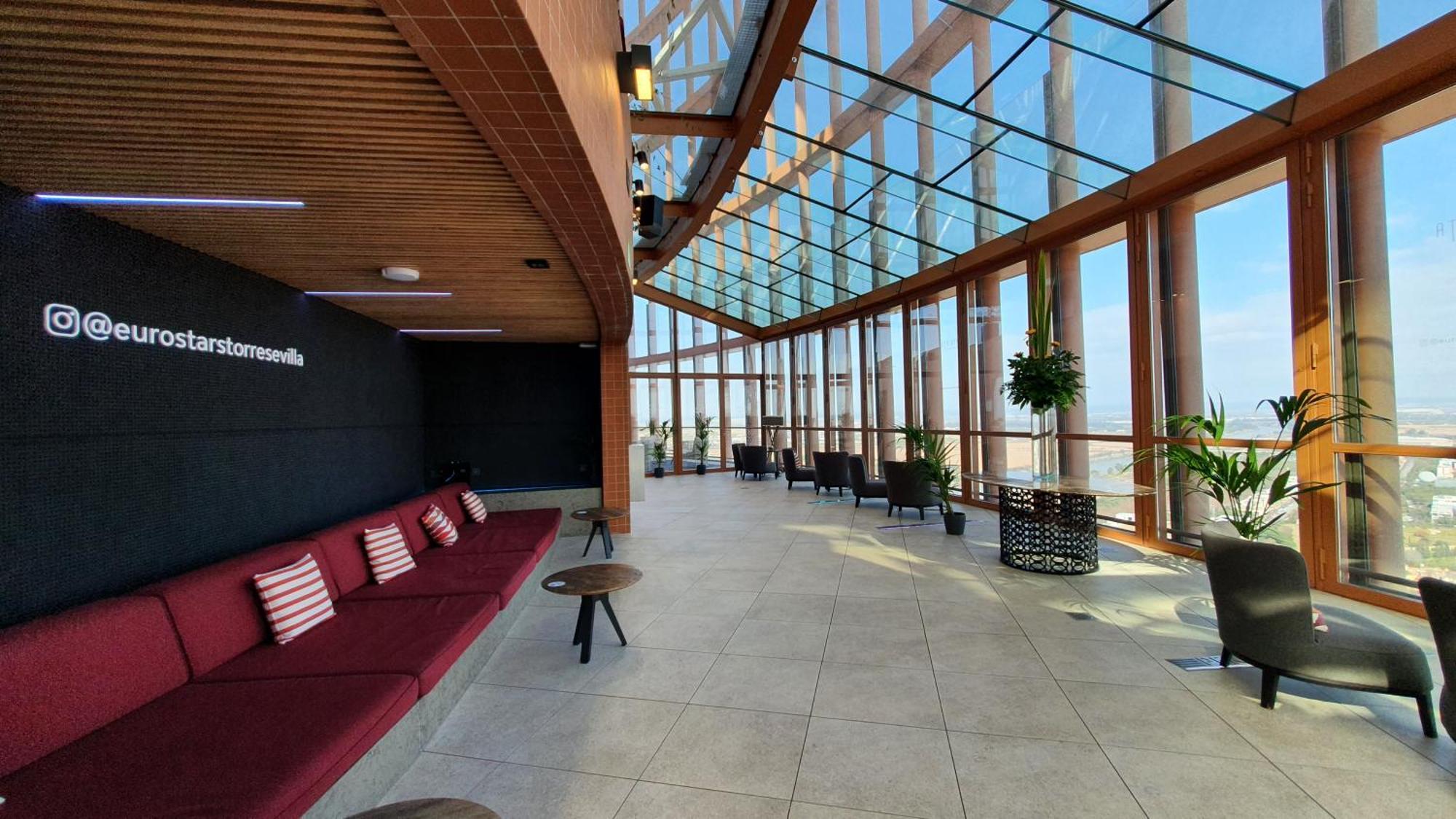 Eurostars Torre Sevilla Esterno foto The photo shows a spacious, modern interior with a large glass wall and ceiling that allows for ample natural light. The area features a long red couch with several cushions, paired with small tables. There are potted plants throughout the space, add