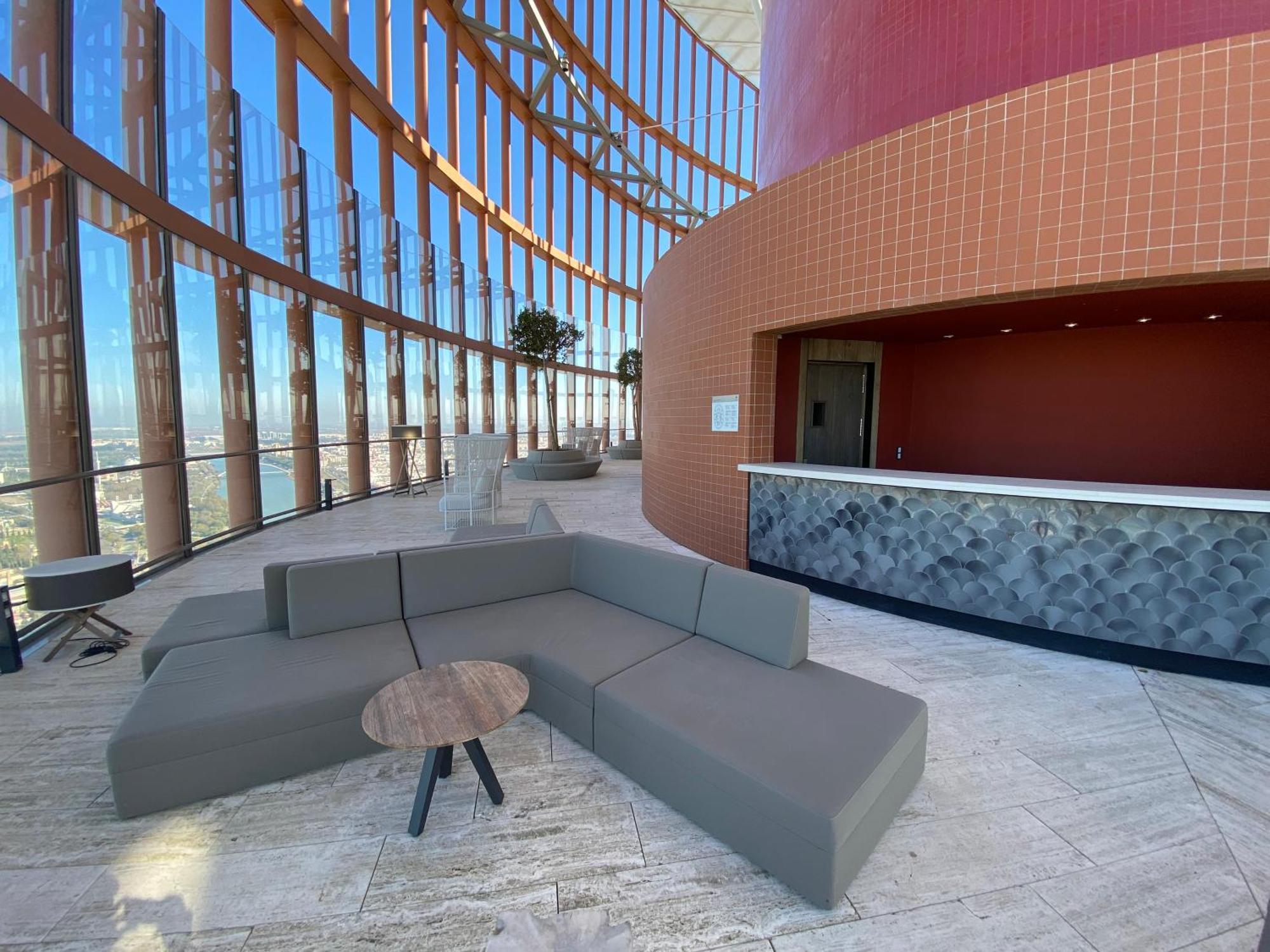 Eurostars Torre Sevilla Esterno foto The photo shows a modern interior space with large windows that provide a panoramic view. There is a comfortable, modular gray seating arrangement shaped like a curved couch, paired with a small, round table. In the background, there's a reception or