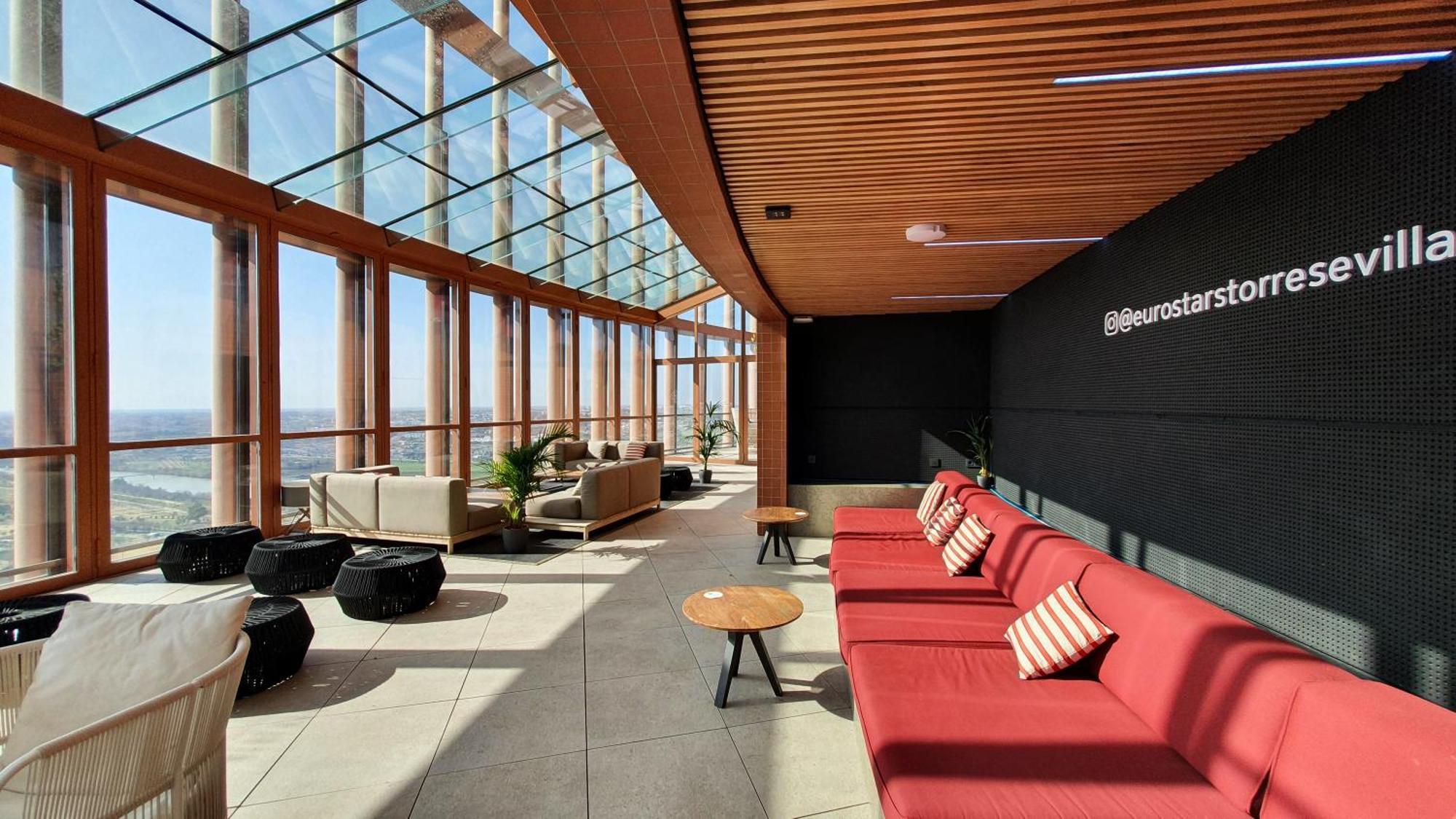Eurostars Torre Sevilla Esterno foto The photo depicts a modern interior space with a spacious, well-lit atmosphere. It features a lounge area that includes several red couches with decorative cushions, arranged in a way to encourage conversation. There are small round tables beside the