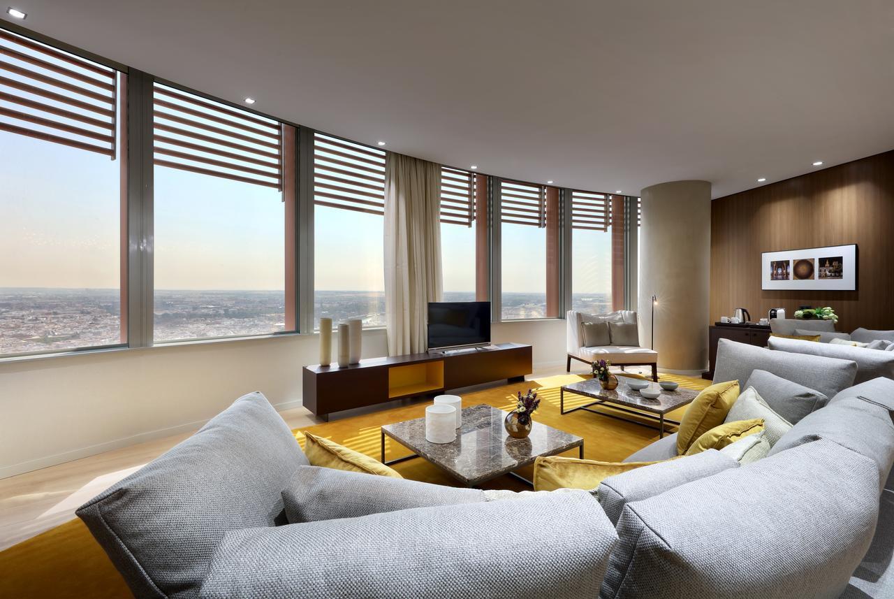 Eurostars Torre Sevilla Esterno foto The photo shows a modern living room featuring large windows that offer a panoramic view. The windows are adorned with horizontal slats that allow for light control. The room is furnished with a stylish, curved sectional sofa in a light color, comple