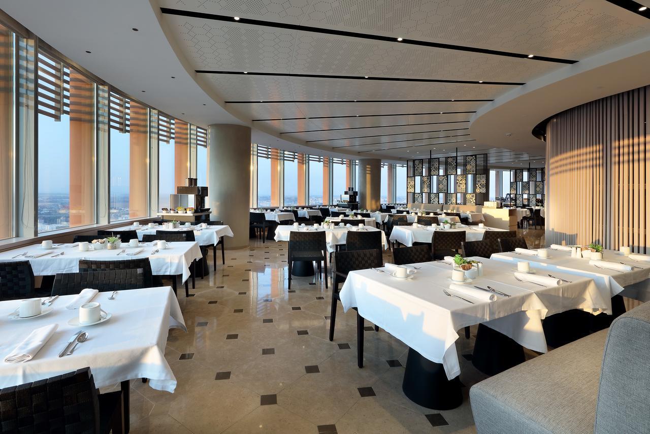 Eurostars Torre Sevilla Esterno foto The photo shows an elegant restaurant interior with a modern design. The space includes several tables, each set with white tablecloths and dishware. The restaurant features large windows that offer a panoramic view, allowing natural light to flood t
