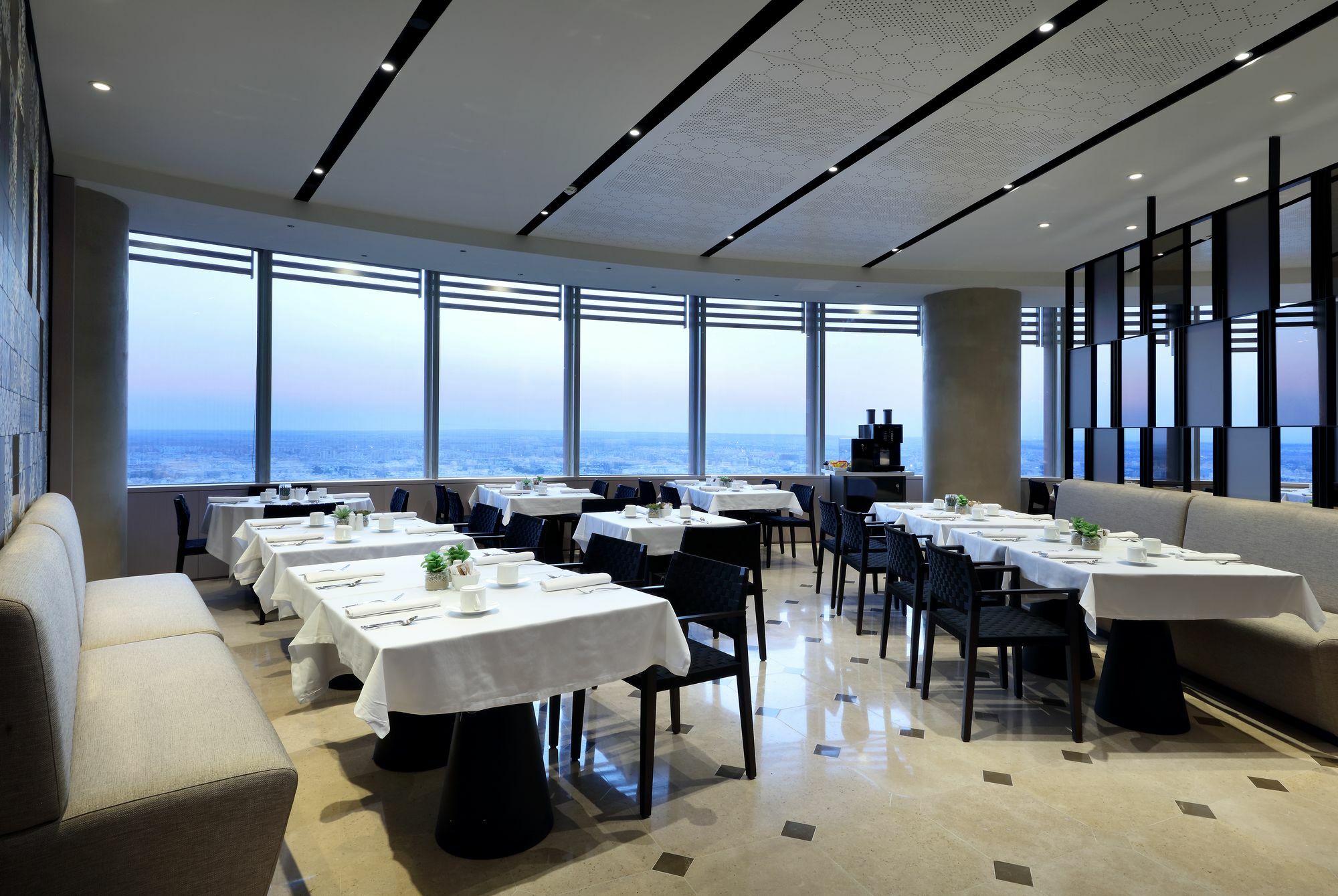 Eurostars Torre Sevilla Esterno foto The photo shows the interior of a modern restaurant. The space features multiple dining tables arranged in an elegant layout, each covered with white tablecloths. The chairs are sleek and dark, complementing the contemporary design. Large windows lin