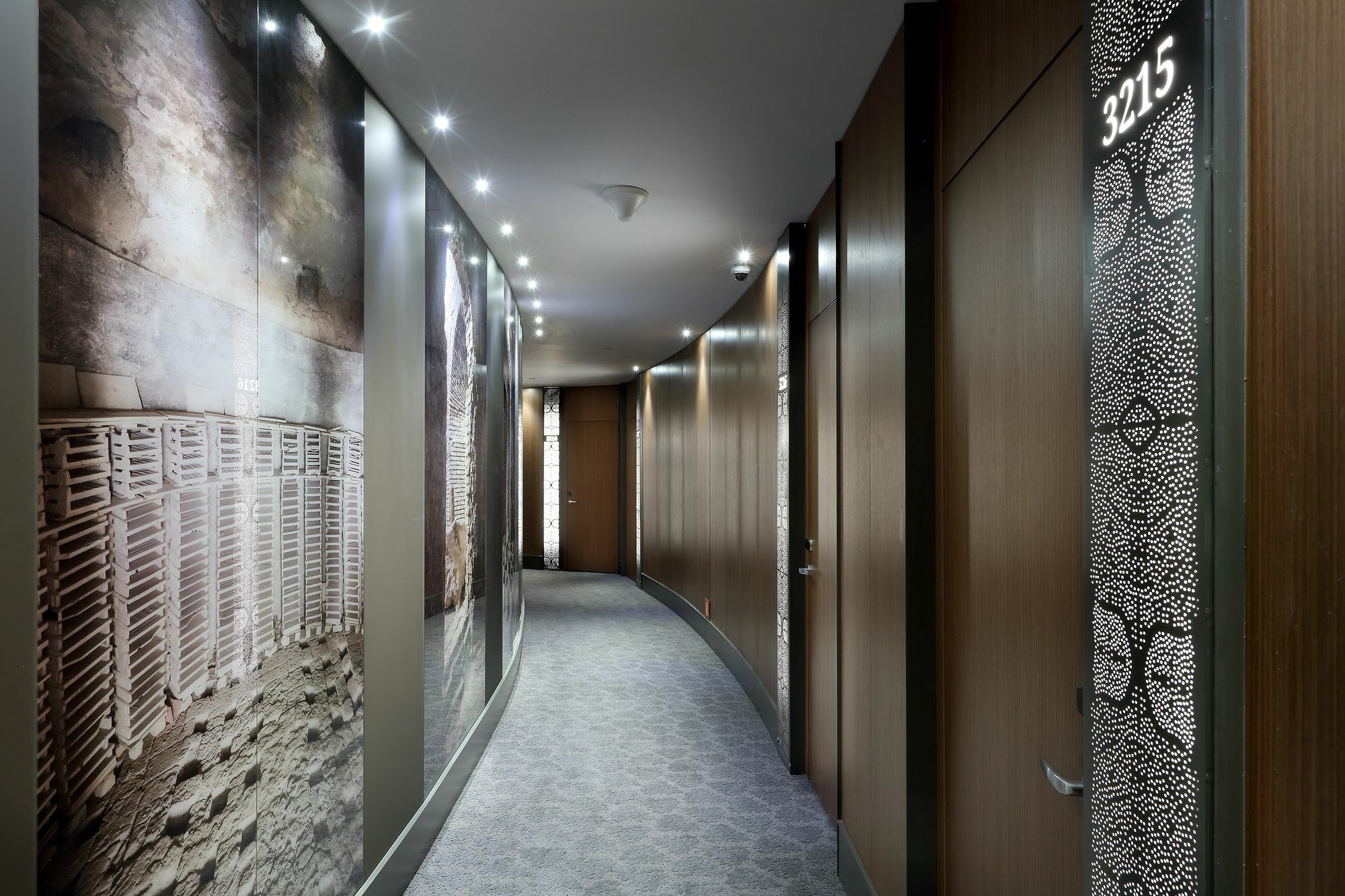 Eurostars Torre Sevilla Esterno foto The photo shows a modern hotel corridor with a curved design. The walls are adorned with large, artistic wall coverings featuring abstract and patterned designs. Soft lighting is embedded in the ceiling, creating a warm ambiance. The doors to guest r