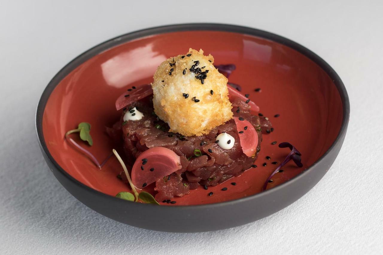 Eurostars Torre Sevilla Esterno foto The photo shows a dish presented in a round, orange bowl. The main component appears to be a tartare, possibly made of raw meat, finely chopped and garnished with small pieces of what looks like radish. Atop the tartare, there's a round, golden-brown